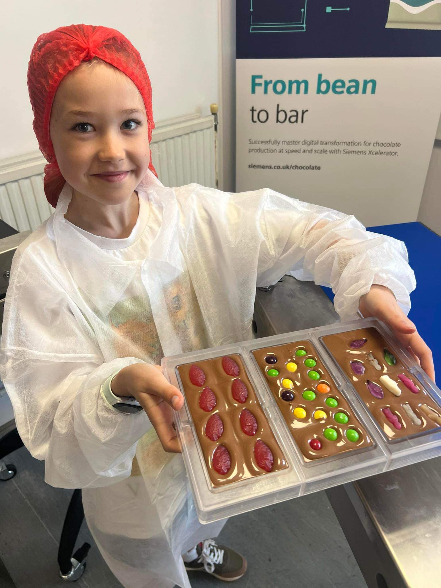 A young chocolate maker in a hairnet and lab coat holding a tray of decorated chocolate bars